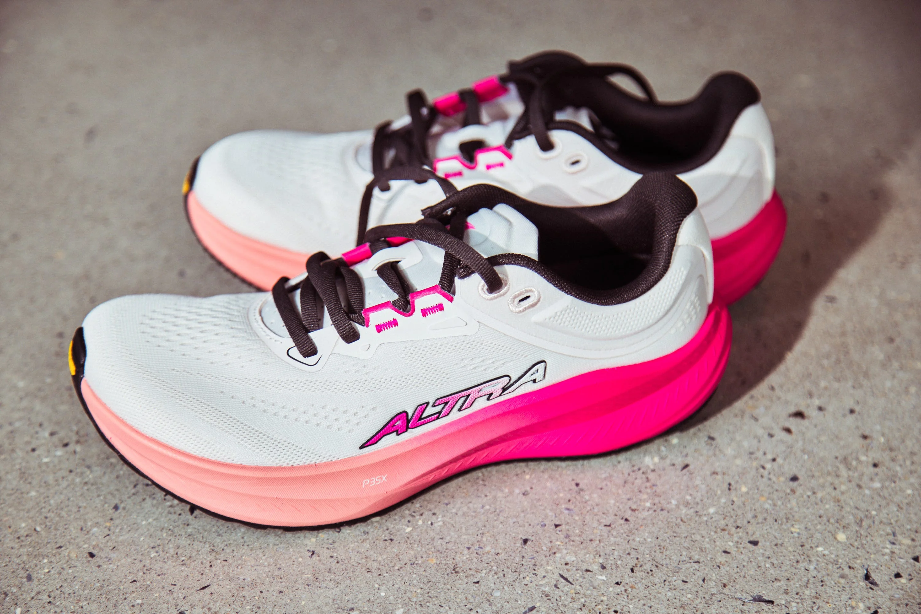 White and pink athletic shoes with black laces and Altra branding on a gray surface. White and pink athletic shoes with black laces and Altra branding on a gray surface.