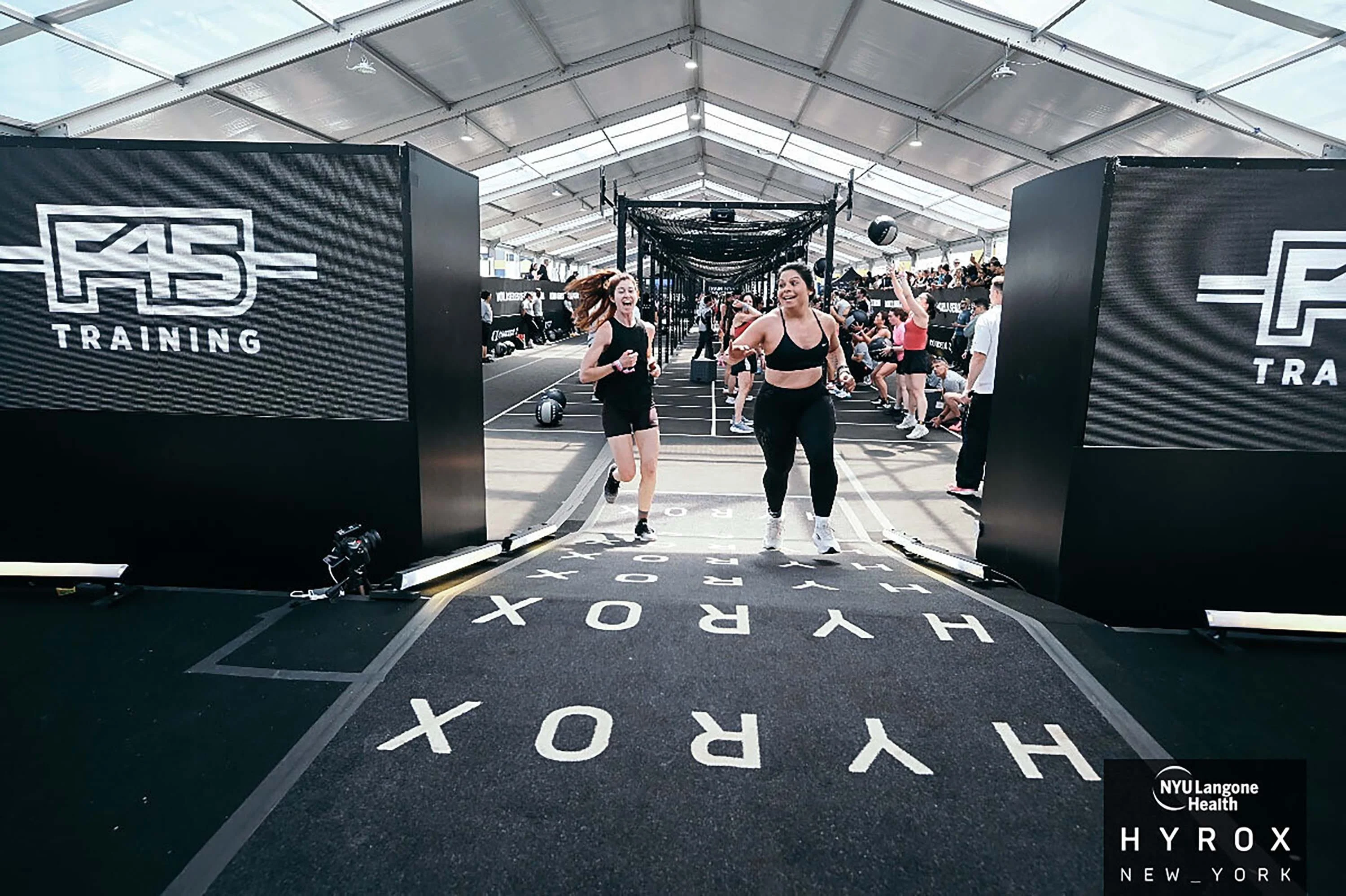Two women running towards the camera in a fitness training event inside a large tent.