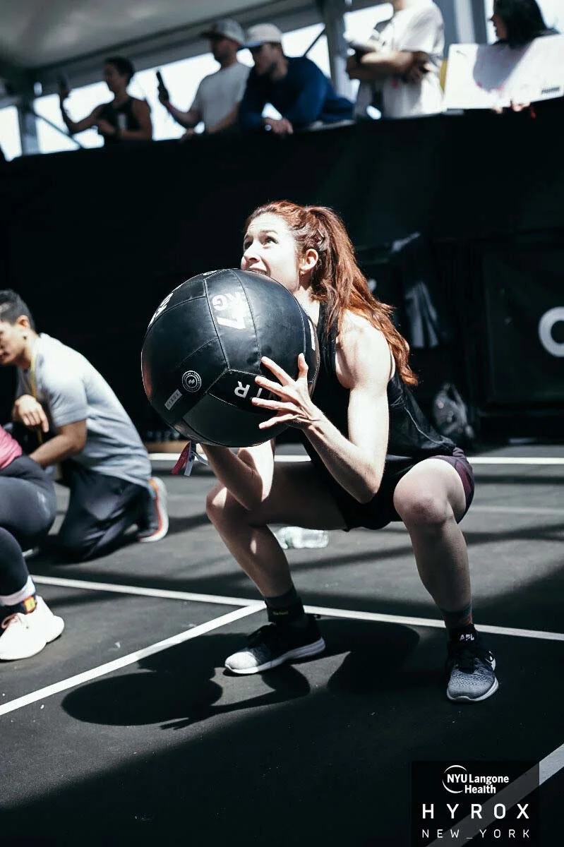 A woman performing a squat while holding a medicine ball during a fitness competition, with spectators in the background.
