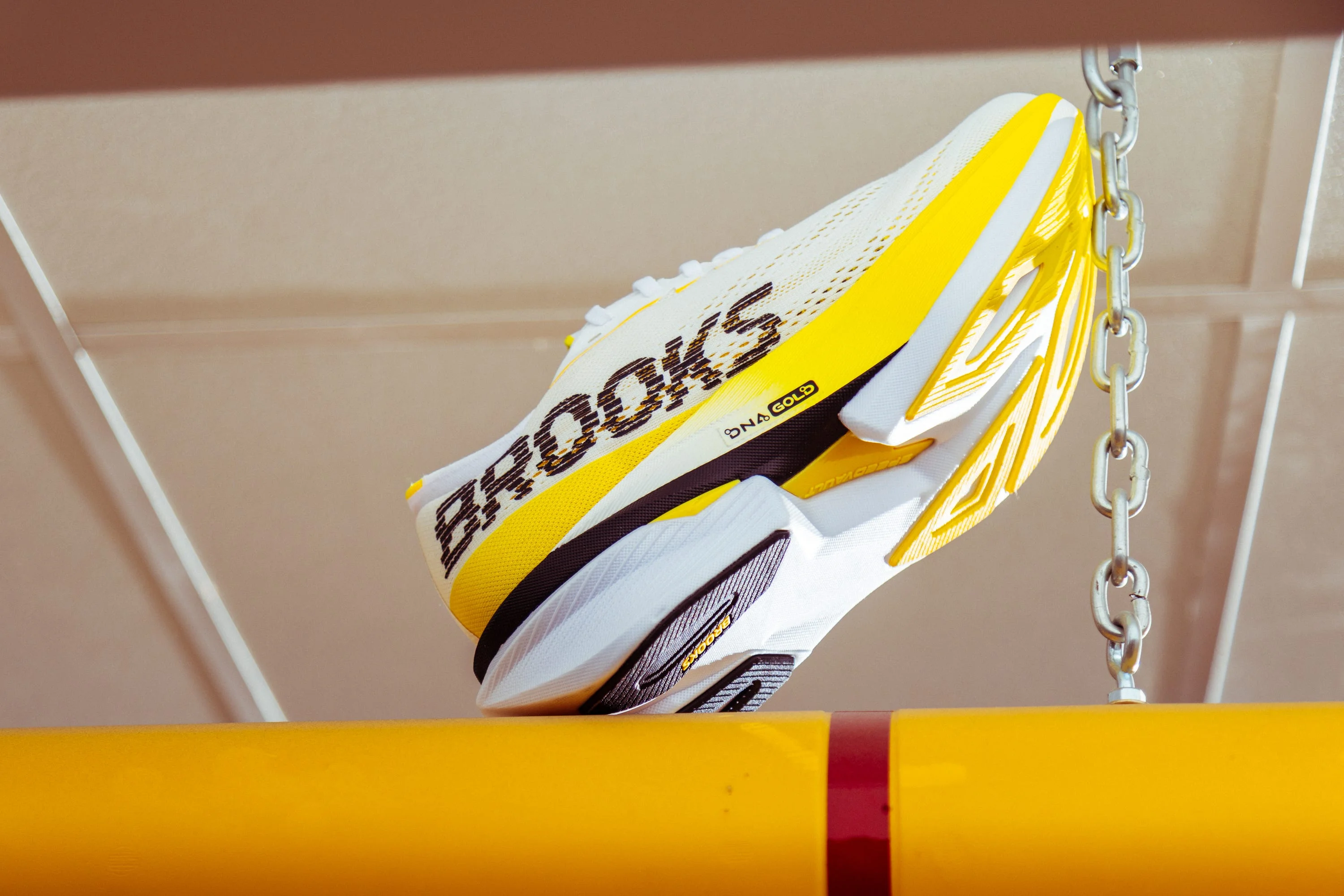 Yellow and white running shoe with "Brooks" branding, displayed hanging above a yellow bar. Yellow and white running shoe with "Brooks" branding, displayed hanging above a yellow bar.