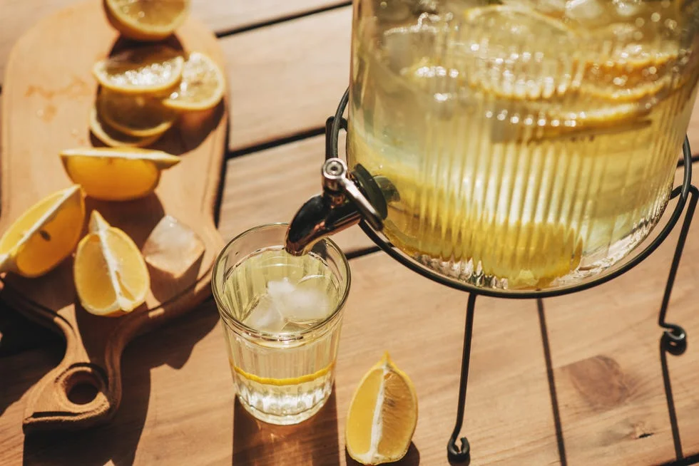 Lemonade with ice in a pitcher and glass on a wooden table with fruit and crushed ice outside with a lemon field in the background on a sunny day. Front view. Horizontal composition. Lemonade with ice in a pitcher and glass on a wooden table with fruit and crushed ice outside with a lemon field in the background on a sunny day. Front view. Horizontal composition.